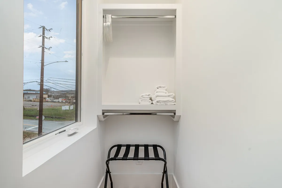 A neatly organized storage area with shelves stacked with towels, a black folding stool, and a large window bringing in natural light. The design is minimal and orderly, offering a sense of calm and readiness.