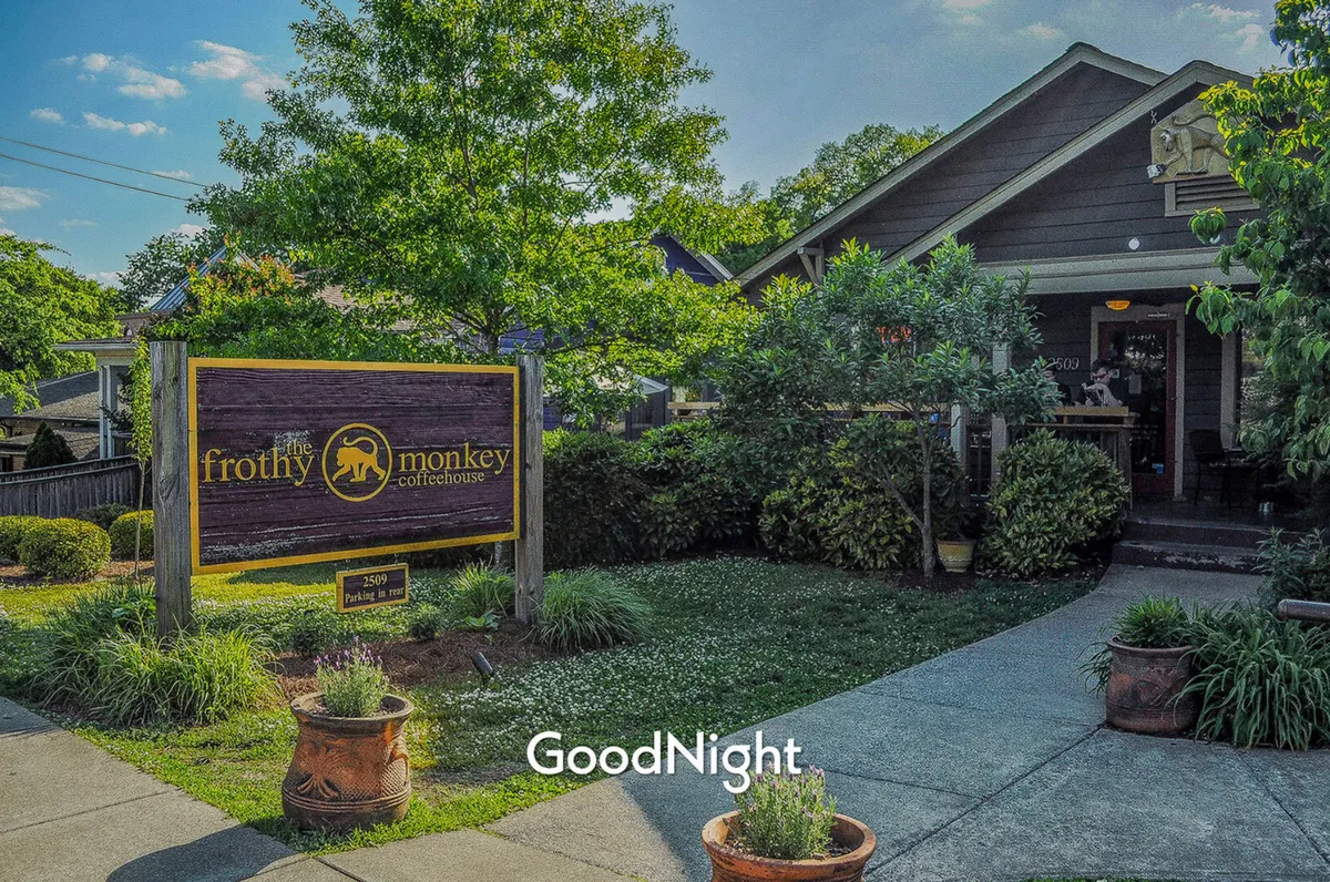 The image captures the exterior of a stylish local café, with prominent signage, neat landscaping, and an inviting building façade. The atmosphere appears friendly and relaxed, suggesting a vibrant gathering spot for coffee and casual conversation just steps from home.