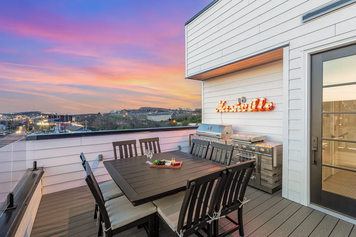 A rooftop dining area equipped with a grill, a stylish outdoor dining set, and a vibrant neon 'Nashville' sign. The sunset sky provides a warm, inviting backdrop, perfect for BBQs and evening gatherings.