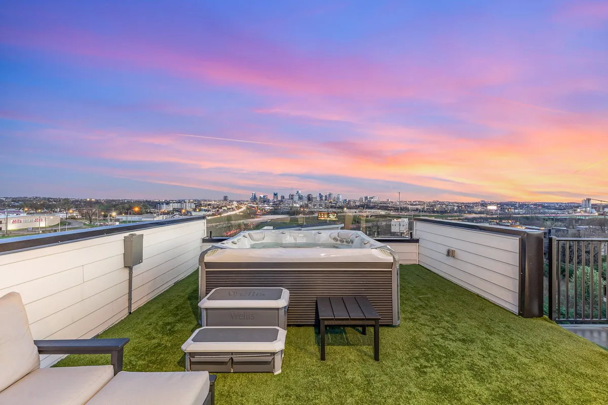 A spacious rooftop deck features a modern hot tub with seating for several people, offering an impressive view of the city skyline. Plush outdoor seating is arranged for groups to relax and socialize. The sun is setting in the background, casting a tranquil glow over the space and enhancing the inviting atmosphere ideal for evening gatherings.