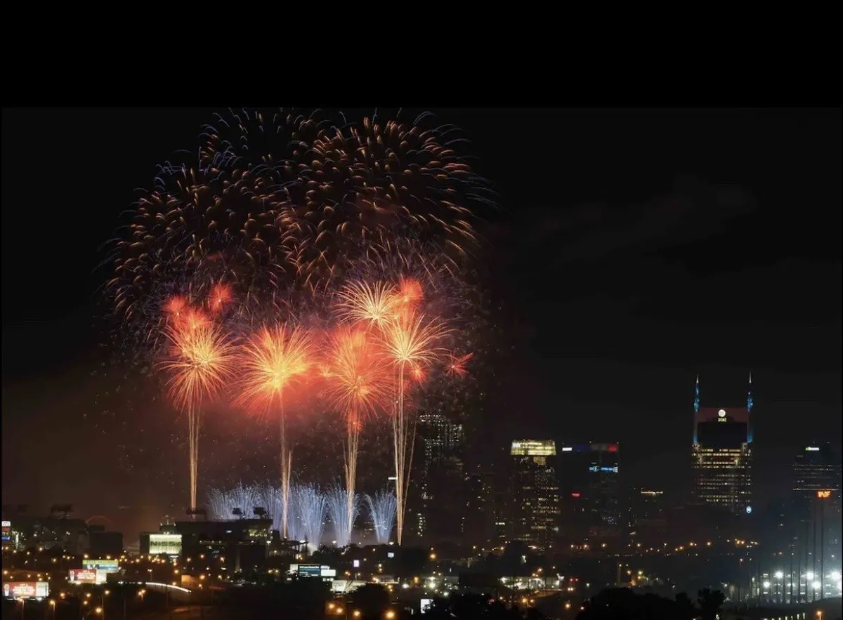 A dazzling fireworks display lights up the night sky above the city skyline, viewed from an elevated outdoor vantage point. Groups of people are visible, sharing a festive moment and enjoying the spectacle together, capturing the essence of special celebrations on the rooftop.