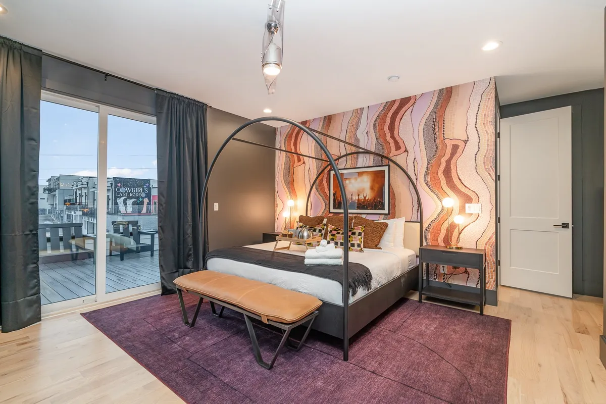 A spacious bedroom featuring a canopy bed, a textured accent wall, large sunlit windows, and curated artwork. The room exudes warmth and an inviting, stylish ambiance, with plush bedding and a comfortable seating area by the window.
