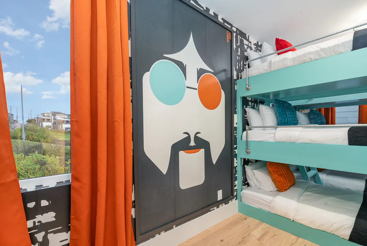 A vibrant and playful bunk room with multiple bunk beds, large wall art of a stylized face with sunglasses, and bright curtains. The layout encourages group stays with ample sleeping accommodations and an artistic flair.