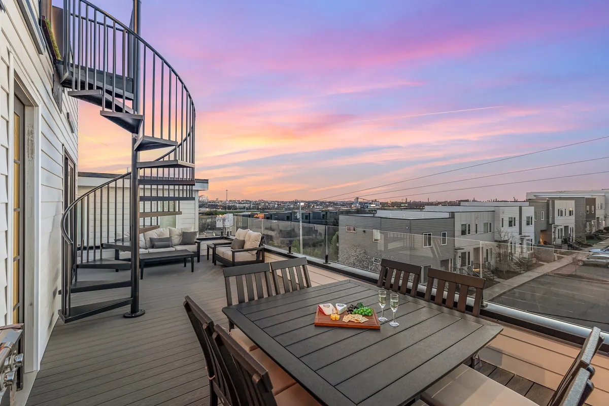 An inviting rooftop terrace with a spiral staircase, a large dining table set for a group, and a vibrant sunset sky. The table is ready for an al fresco meal, and the scene suggests a lively yet relaxed gathering space ideal for celebrations, BBQ dinners, and enjoying panoramic views.