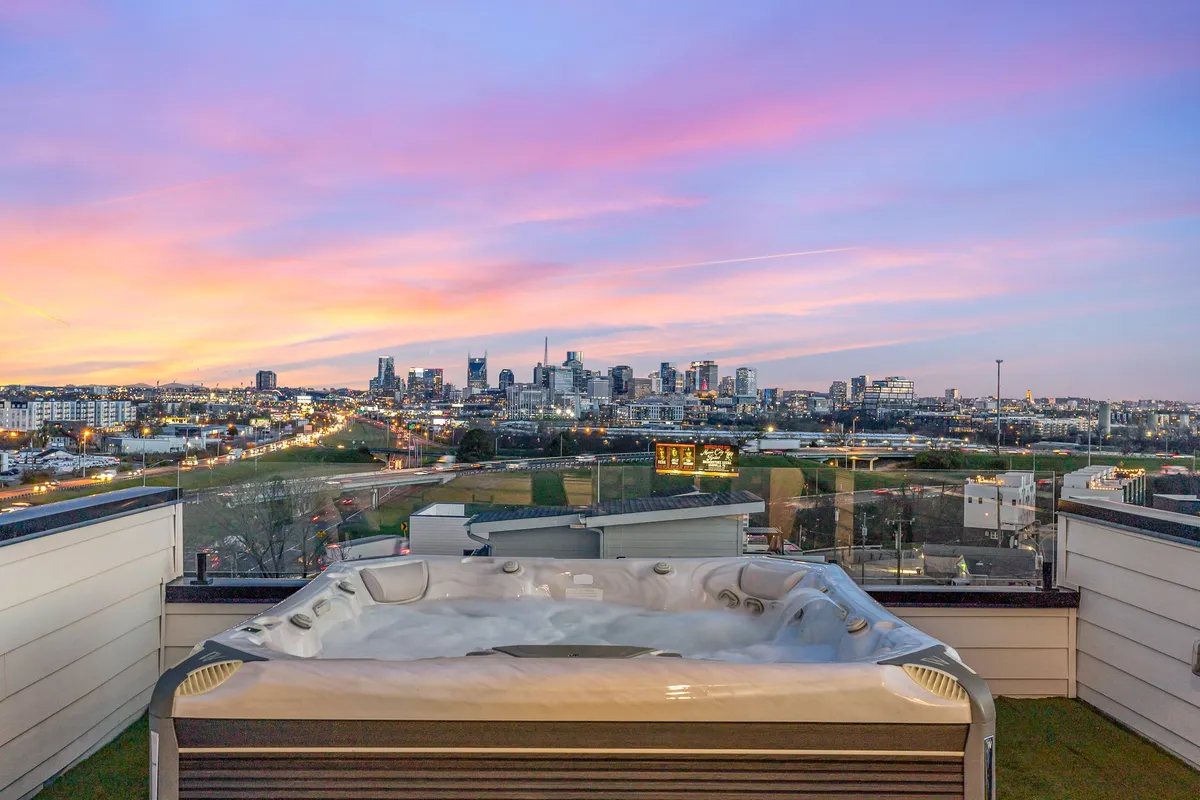 The image captures a stunning rooftop deck at sunset, with a luxurious hot tub in the foreground. The Nashville city skyline glows in the distance under vibrant sunset colors, creating a romantic and tranquil atmosphere perfect for relaxation or entertaining friends.